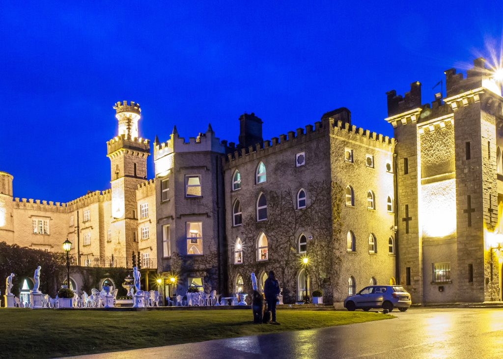 A majestic view of Cabra Castle surrounded by lush green lawns and woodlands, with its stone towers and battlements under a soft, overcast sky.
