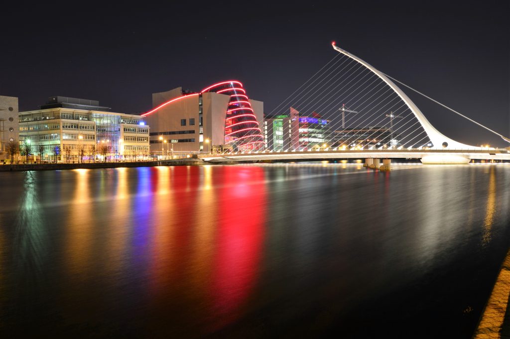 A panoramic view of Dublin city with the River Liffey flowing through the center, bordered by historic buildings and modern architecture under a cloudy sky.
